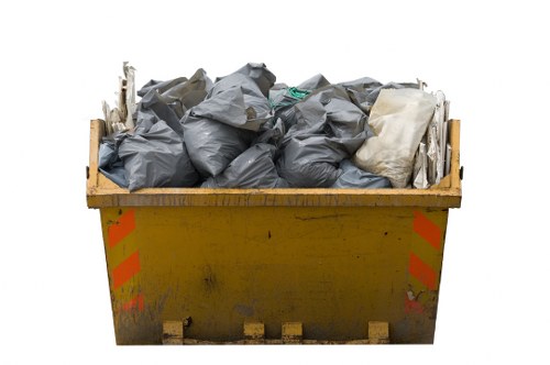 A large yellow industrial skip filled with tightly packed black rubbish bags and some light-colored bags, situated on a paved or concrete surface. The skip appears to be outdoors, with some visible dirt and minor graffiti or stains on its sides. The bags are piled up to the top of the skip, which is designed for waste collection and disposal, commonly used by commercial waste removal services such as Commercial Waste Removal Walthamstow. The image focuses solely on the skip and its contents, with no other objects or background elements visible, emphasizing the collection of waste materials ready for removal or recycling. The environment appears functional and utilitarian, typical for a waste disposal setting, aligning with the company's rubbish removal services in the Walthamstow area of London, E17 postcode.
