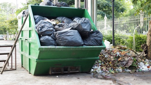 A large green Skip bin filled with tightly packed black rubbish bags sits on a paved outdoor area, likely on a driveway or street in Walthamstow. The bin has a sturdy metal construction with visible textured surfaces and is positioned beside a pile of mixed waste, including paper, plastic, and other debris, which is scattered on the ground near the bin. In the background, there are green trees, a metal fence, and some foliage, indicating a residential or park setting. The scene is illuminated by natural daylight, highlighting the contrast between the dark rubbish bags and the bright green of the bin. This image relates to waste management and rubbish removal services provided by Commercial Waste Removal Walthamstow, emphasizing the collection and disposal of household or commercial waste in the area.
